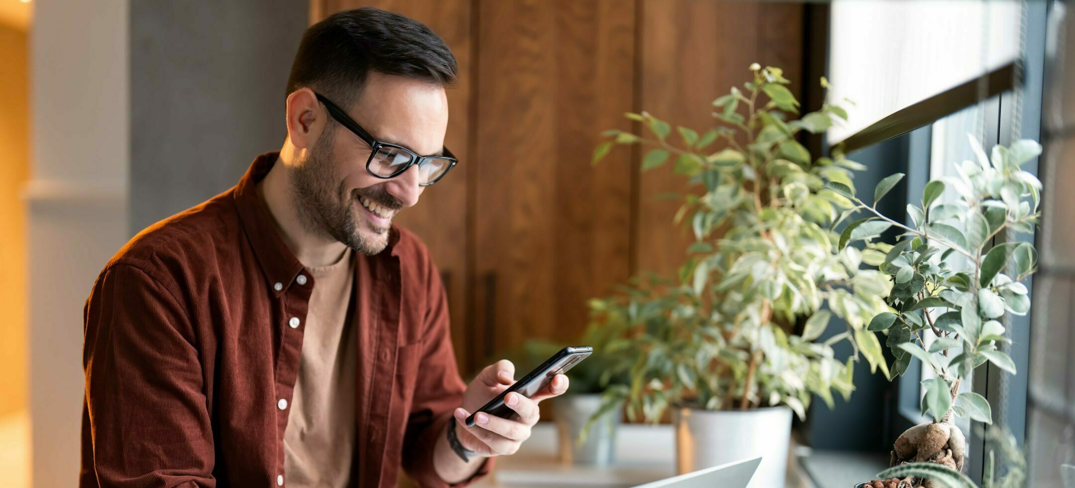 Satisfied modern millennial man in stylish casual clothes using smartphone and laptop computer for electronic banking, making reservation, online shopping and payments while spending time at home.